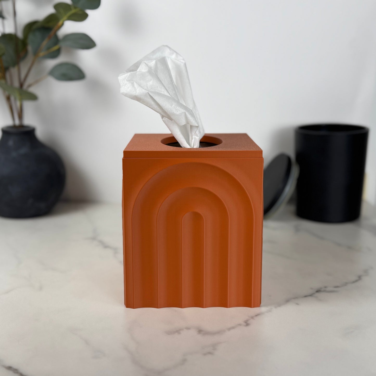 Terracotta colored tissue box with tissues on a marble surface with decorative items in the background.