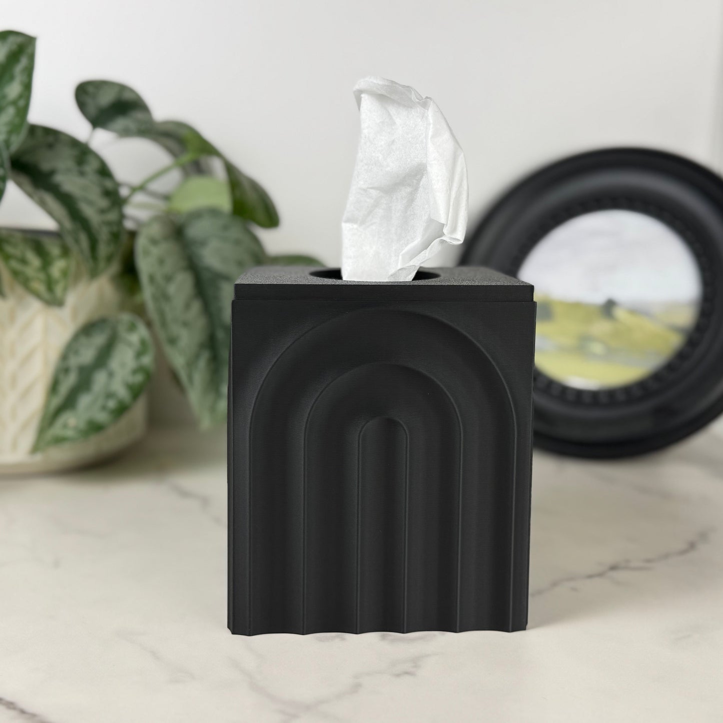 Black tissue box holder on a marble surface with a plant and decorative item in the background