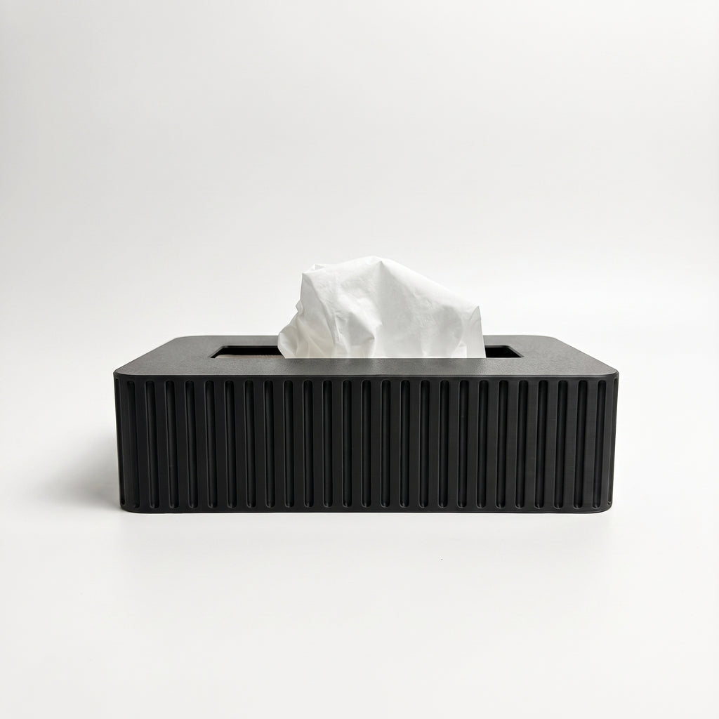 A rectangular tissue box cover in a black color with vertical fluted ridges, shown with a white tissue protruding from the top on a clean white background.