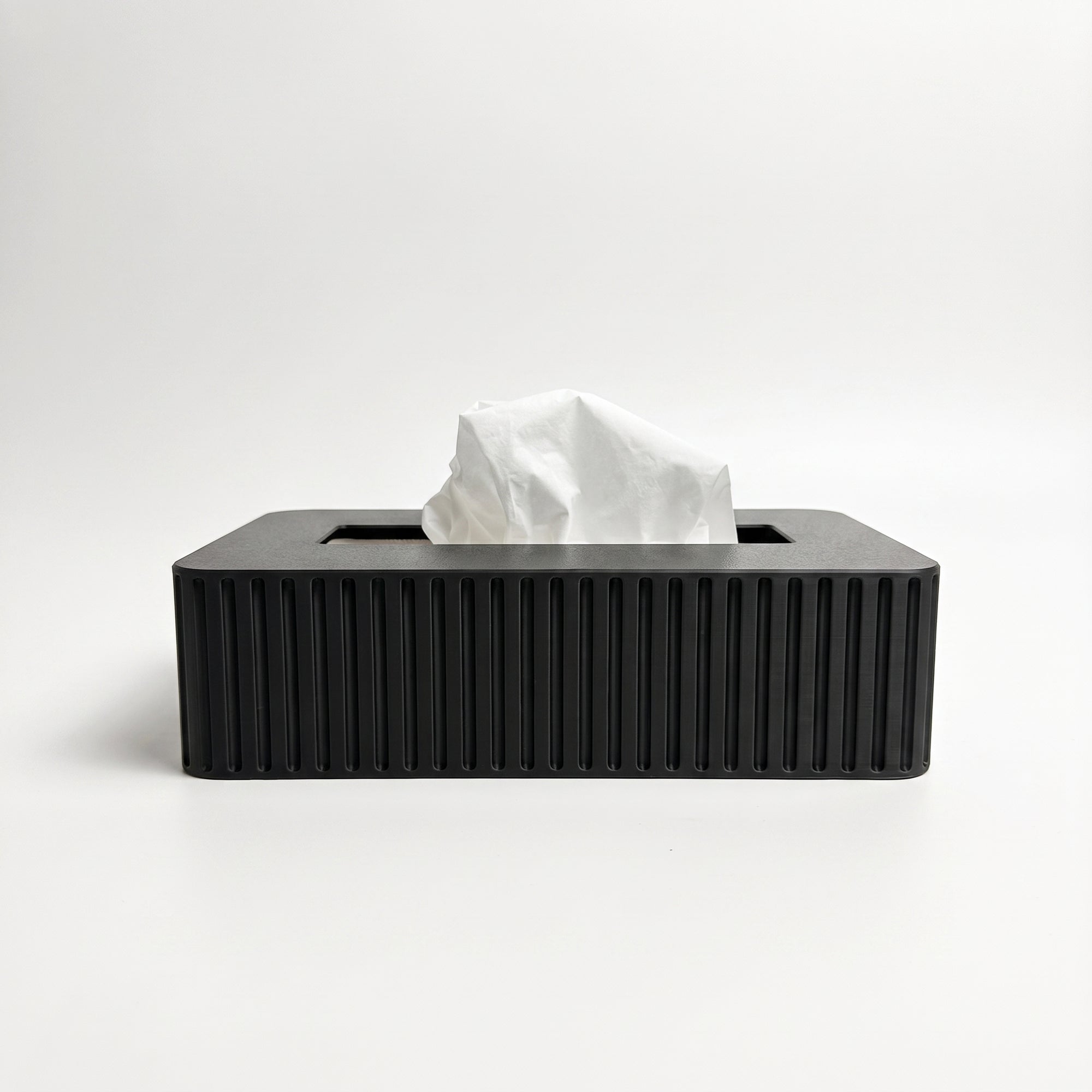 A rectangular tissue box cover in a black color with vertical fluted ridges, shown with a white tissue protruding from the top on a clean white background.